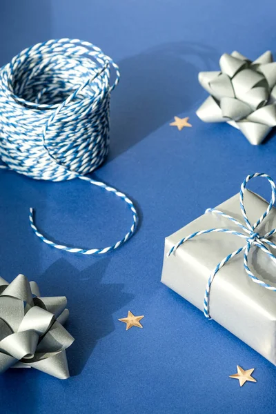 Gift packaging: silver bows for gift decoration, blue gift rope, gift in a silver box on a blue background. Preparing for Christmas holidays. Selective focus