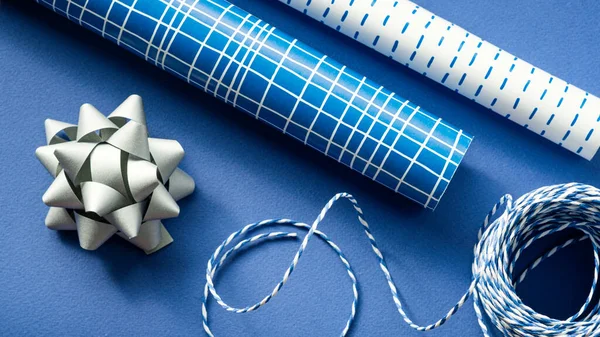 Rolls of Christmas wrapping paper, silver bows and blue gift rope on a blue background. Preparing for Christmas holidays. Gift packaging.