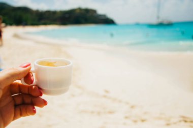 Antigua 'da bir plajda otururken ve manzaranın tadını çıkarırken elinde küçük bir fincan kahve tutuyor. Metin uzayı. Yatay
