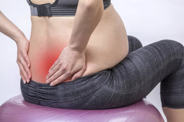 La mujer embarazada activa hace ejercicio en el gimnasio en la sala de yoga en la pelota de fitness y se toma de la mano en la espalda para aliviar el dolor de espalda — Foto de Stock