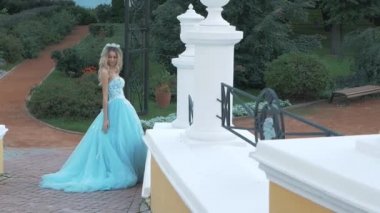 Sevimli sarışın kız mavi bir elbise sarayda dışarı kamera Bahçe kendi bakar gelir ve yeşilliklerle görünümünü sahiptir, kamerada poz veriyor, elleri vuruş