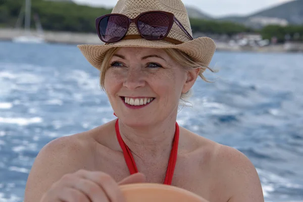 Croatia, Hvar - June 2018: Women in mid 40's driving a small boat.  She wears a red dress and hat hat and sunglasses on her head. Coastline is visible in the background.