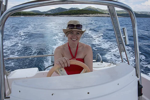 Croatia, Hvar - June 2018: Women in mid 40's driving a small boat.  She wears a red dress and hat hat and sunglasses on her head. Coastline is visible in the background.