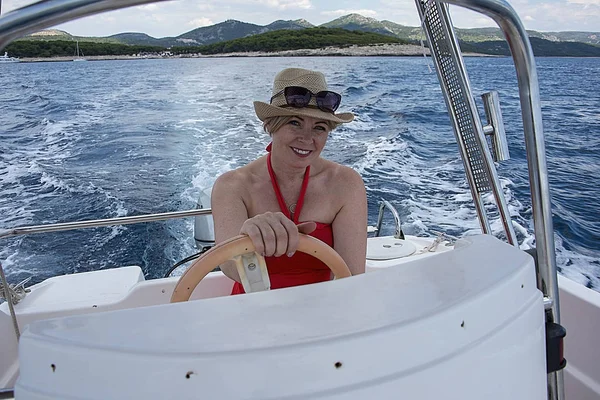 Croatia, Hvar - June 2018: Women in mid 40\'s driving a small boat.  She wears a red dress and hat hat and sunglasses on her head. Coastline is visible in the background.