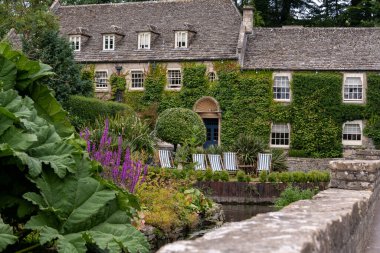 Bibury, İngiltere, İngiltere - 10 Eylül 2021: Renkli çiçeklerle kaplı sarmaşık kaplı taş bir evin önünde çizgili bahçe sandalyeleri duruyor