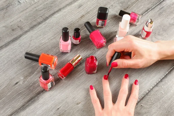 Top view on young woman hands, applying second red coat of nail varnish, with glass bottles with nail polish in the background, on gray wood desk.