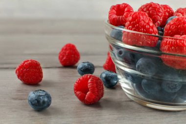 Yakın, cam kase yaban mersini tam üstünde bir kýsmýný ahududu ile gri ahşap masanın üstüne dökülen.