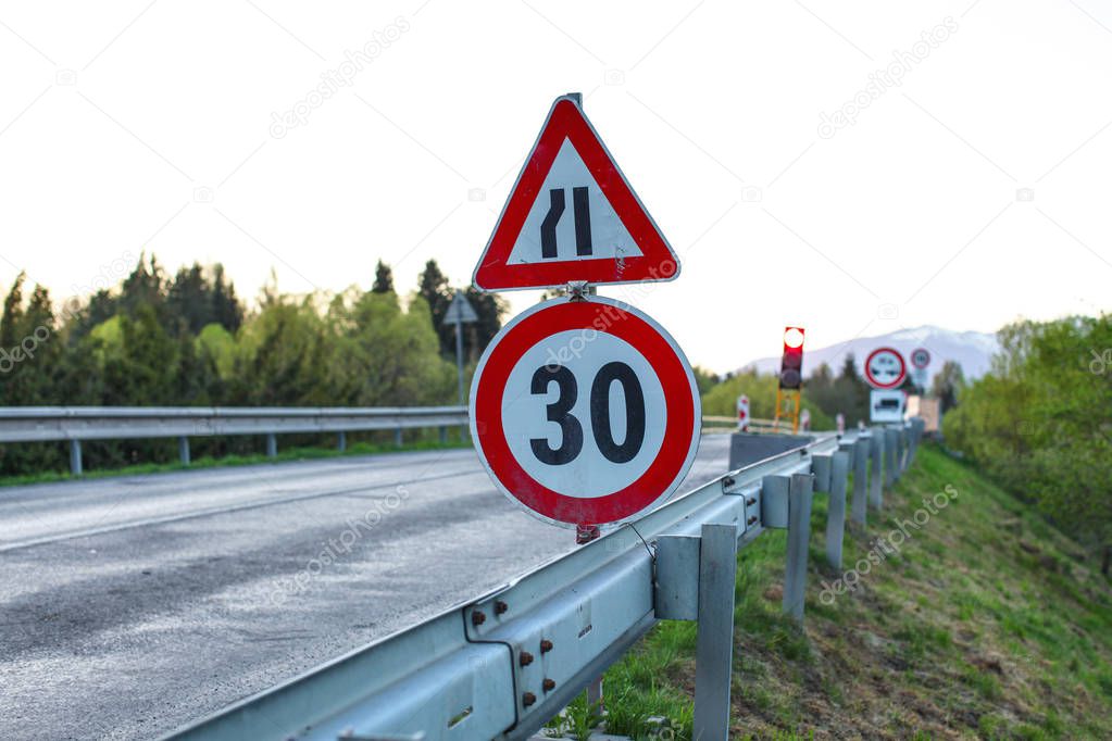Road sign, reduce speed to 30, blurred red traffic light and construction in background, shot on summer evening.