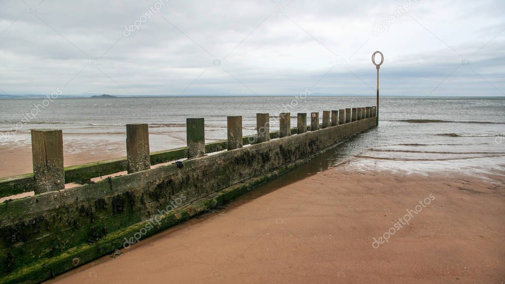 Old wooden and stone groyne structure covered with green algae on Portobello beach, low tide North sea in the backgroud, on overcast day. Edinburgh, Scotland.
