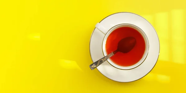 Asztali nézet, süt a nap reggel fehér porcelán csésze forró tea, ezüst kanállal, a sárga board - széles fotó tér bal oldalán szöveg — Stock Fotó