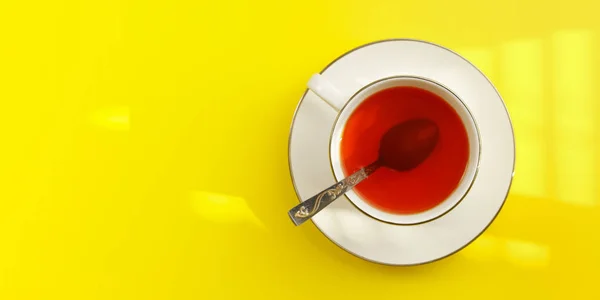 Tabletop view, morning sun shines to white porcelain cup with hot tea, silver spoon in, on yellow board - wide photo space for text left side — Stock Photo, Image