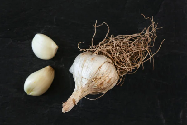 Vista de arriba hacia abajo, bulbo de ajo natural con raíces, dos dientes pelados, en tablero negro —  Fotos de Stock