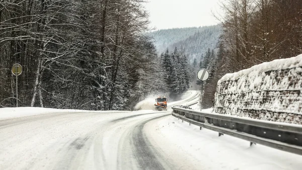 Route forestière enneigée, glace sur asphalte, camion de charrue à râpe orange venant au loin, conditions de conduite dangereuses — Photo