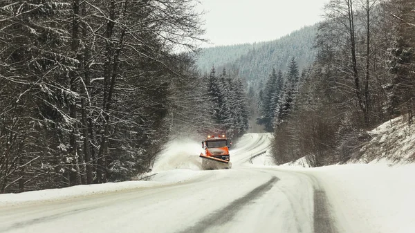 Bright orange plough truck cleaning snow on forest road in winter, view from car driving other way, trees around, overcast day