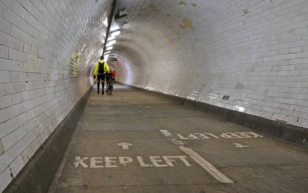 Ciclista con giacca gialla che cammina con la bicicletta all'interno del tunnel pedonale di Greenwich sotto il Tamigi, pedoni più sfocati sullo sfondo, tenere il cartello a sinistra sul pavimento — Foto Stock