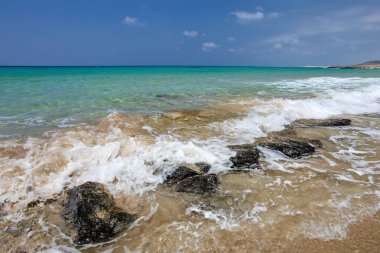 Küçük deniz dalgaları kumlu sahilde siyah kayalara çarpıyor.