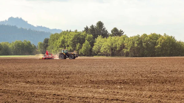 Trattore semina in campo vuoto sulla campagna, piccoli alberi in bac — Foto Stock