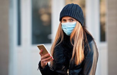 Siyah ceketli ve bereli genç bir kadın. Tek kullanımlık mavi virüs maskesi takıyor. Cep telefonunu kontrol ediyor. Coronavirus covid-19 salgın önleme kavramı