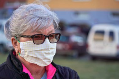 Ev yapımı giysiler giyen yaşlı bir kadın dışarıda ağız virüsü maskesi, binalar ve arabalar bulanık. Coronavirus covid-19 salgını önleme sırasında kullanılabilir