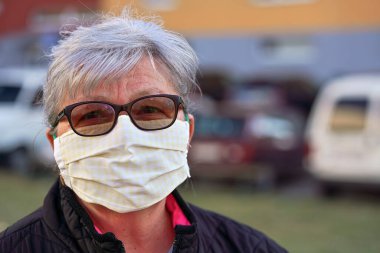 Ev yapımı giysiler giyen yaşlı bir kadın dışarıda ağız virüsü maskesi, binalar ve arabalar bulanık. Coronavirus covid-19 salgını önleme sırasında kullanılabilir