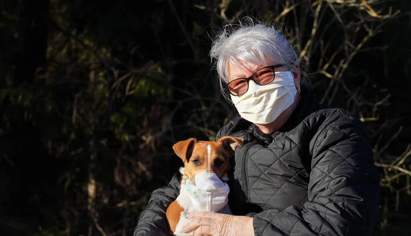 Senior elderly woman wearing virus mask sitting in park with her dog wearing simple respirator as well. Pets are not vulnerable to coronavirus covid-19 but some owners protect them nevertheless