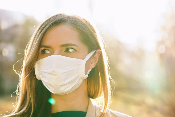 Young woman wearing white cotton virus mouth nose mask, nice backlight sun bokeh in background, closeup face portrait