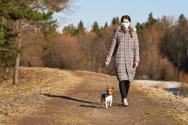 Sıcak ceketli, virüs suratlı, ağız maskesi takan, kır yolunda köpeğini gezdiren genç bir kadın. Bazı ülkelerde Coronavirus covid-19 salgını sırasında maskeler evin dışında zorunludur.