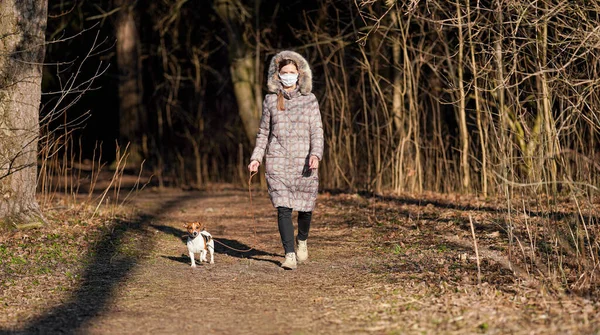 Young woman in warm jacket, wearing virus face mouth mask, walks her dog on country park road. Masks are mandatory  outside home during coronavirus covid-19 outbreak in some countries
