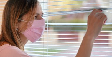 Pembe evdeki genç kadın dışarıdaki pencere perdelerinden bakarak pamuk virüsü maskesi yaptı. Karantina ya da Coronavirus covid-19 salgını sırasında evde kalmak
