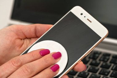 Kadın cep telefonunu pamuklu kağıtla temizliyor, siyah ekranı siliyor, yakın plan detaylarıyla. Arka planda bulanık dizüstü bilgisayar klavyesi. Coronavirus covid-19 salgını önleme