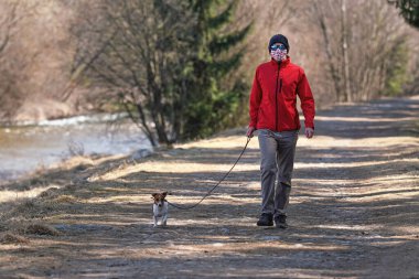 Kırmızı ceketli, bereli ve güneş gözlüklü, ev yapımı virüs maskeli, kır yolunda köpek gezdiren bir genç. Bazı ülkelerde Coronavirus covid-19 salgını sırasında maskeler evin dışında zorunludur.