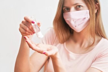 Evinde pembe pamuklu, ağız virüsü maskesi takan, eline anti bakteriyel alkol sürme jeli süren, arka planda bulanık bir yüzü olan genç bir kadın. Coronavirus covid-19 önleme