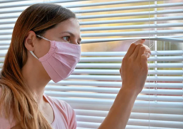 ピンクの家の若い女性は 窓のブラインドを外から見て コットンウイルスの顔マスクを作りました コロナウイルスCovid 19発生中に自宅に隔離または滞在 — ストック写真