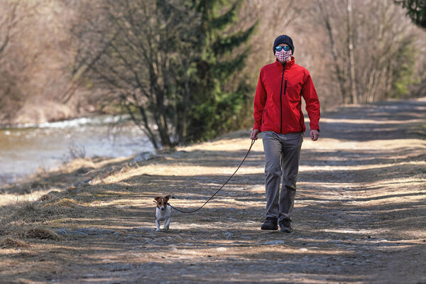 Молодой человек в красной куртке, шапочке и солнцезащитных очках, в домашней вирусной маске, выгуливает собаку по проселочной дороге. Маски являются обязательными вне дома во время вспышки коронавируса ковид-19 в некоторых странах
