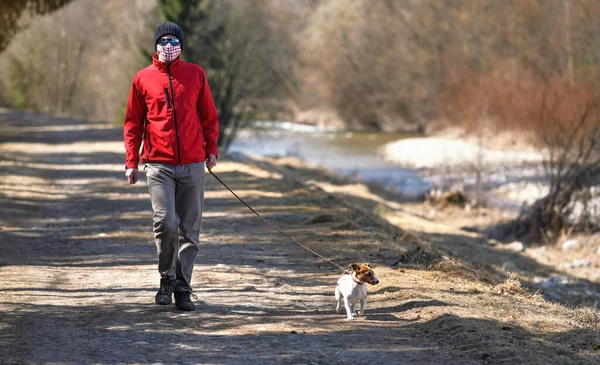 Man in red jacket and white cotton mouth virus mask walks his dog on country road near river. Masks are mandatory   outside home during coronavirus covid-19 outbreak in some countries