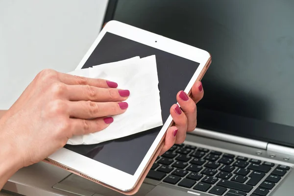 Woman cleaning tablet device with cotton paper tissue, wiping black screen, closeup detail pink color fingernails. Blurred laptop keyboard background.