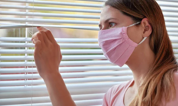 Young woman in pink home made cotton virus face mask, looking through window blinds outside. Quarantine or stay at home during coronavirus covid-19 outbreak