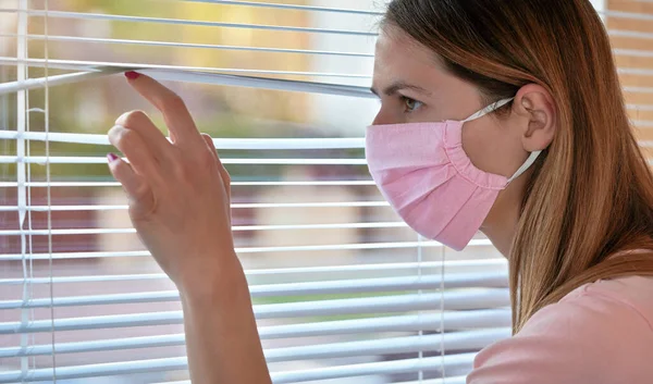ピンクの家の若い女性は 窓のブラインドを外から見て コットンウイルスの顔マスクを作りました コロナウイルスCovid 19発生中に自宅に隔離または滞在 — ストック写真