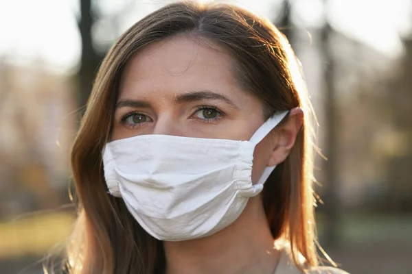 Young woman wearing white cotton virus mouth nose mask, blurred park and trees background, closeup face portrait