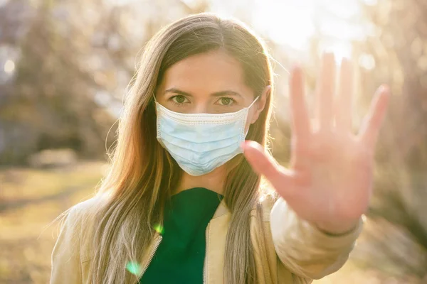 Young woman wearing blue disposable single use virus mouth nose mask, hold hand front of her as stop gesture, blurred sunset lit trees in background. Coronavirus covid 19 outbreak prevention concept