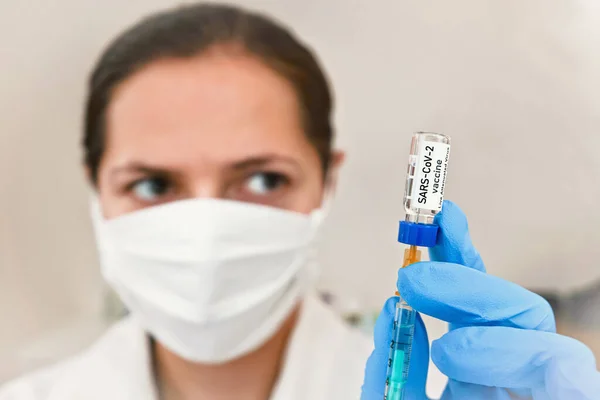 青い手袋で手で注射器に注射されたコロナウイルスワクチン ラベル自身のデザインではなく 実際の製品 白い綿マスクの背景にぼやけた看護師の顔 — ストック写真