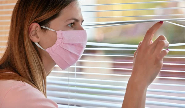 Young woman in pink home made cotton virus face mask, looking through window blinds outside. Quarantine or stay at home during coronavirus covid 19 outbreak