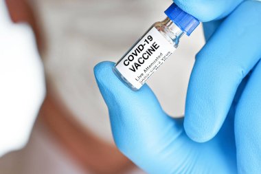Üzerinde Covid 19 aşı etiketi olan küçük bir şişe tutan mavi tıbbi eldivenler (çıkartma gerçek ürün değil, kendi tasarımı), pamuk maskeli bulanık bir yüz. Coronavirus tedavisi kavramı