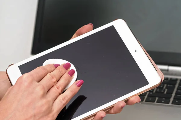 Woman cleaning tablet device with cotton paper tissue, wiping black screen, closeup detail. Blurred laptop keyboard background