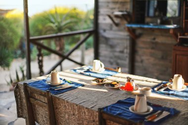 Sabah güneşi masada boş tabaklar ve kahve fincanları ile parlıyor, ortada cam çay demliği, sabah kahvaltısı için hazır, arka planda bulanık yeşil yapraklar - tropikal tatil beldesi