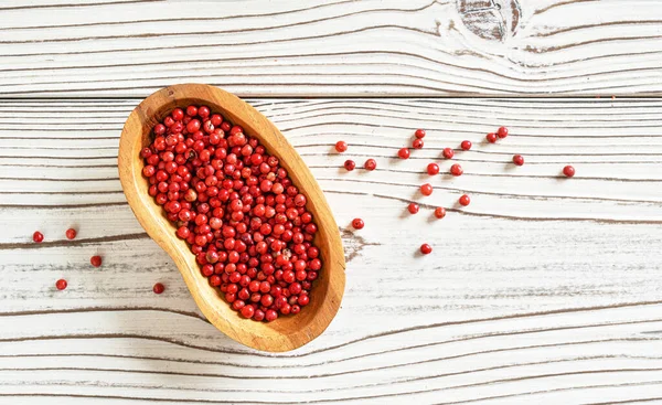 Red or Pink peppercorn in small wooden bowl, some scattered on white boards desk, closeup photo from above