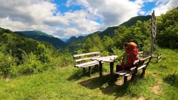 若い登山家の女性 バックパックで彼女の背中に休憩所で素朴なテーブルで山の景色を楽しんで 晴れた夏の日 — ストック動画