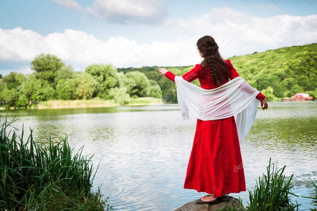 girl in a red historical dress stands on the banks of the river