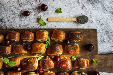 Kirazlı haşhaşlı çörekler, nane yaprakları ve kahverengi tahta üzerinde haşhaş tohumları olan bir kaşık.