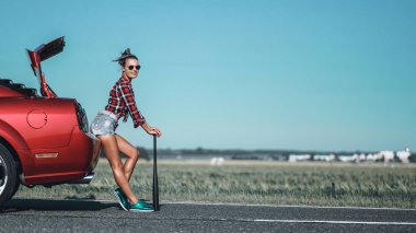 elinde bir beyzbol yarasa ile güzel esmer kız kısa şort giyiyor ve kırmızı spor araba yakın güneş gözlüğü ile bir gömlek